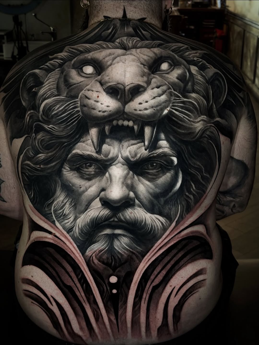 Stacked lion and bearded man portrait tattoo on chest with flowing pink decorative lines below, rendered in black and grey with smooth shading.