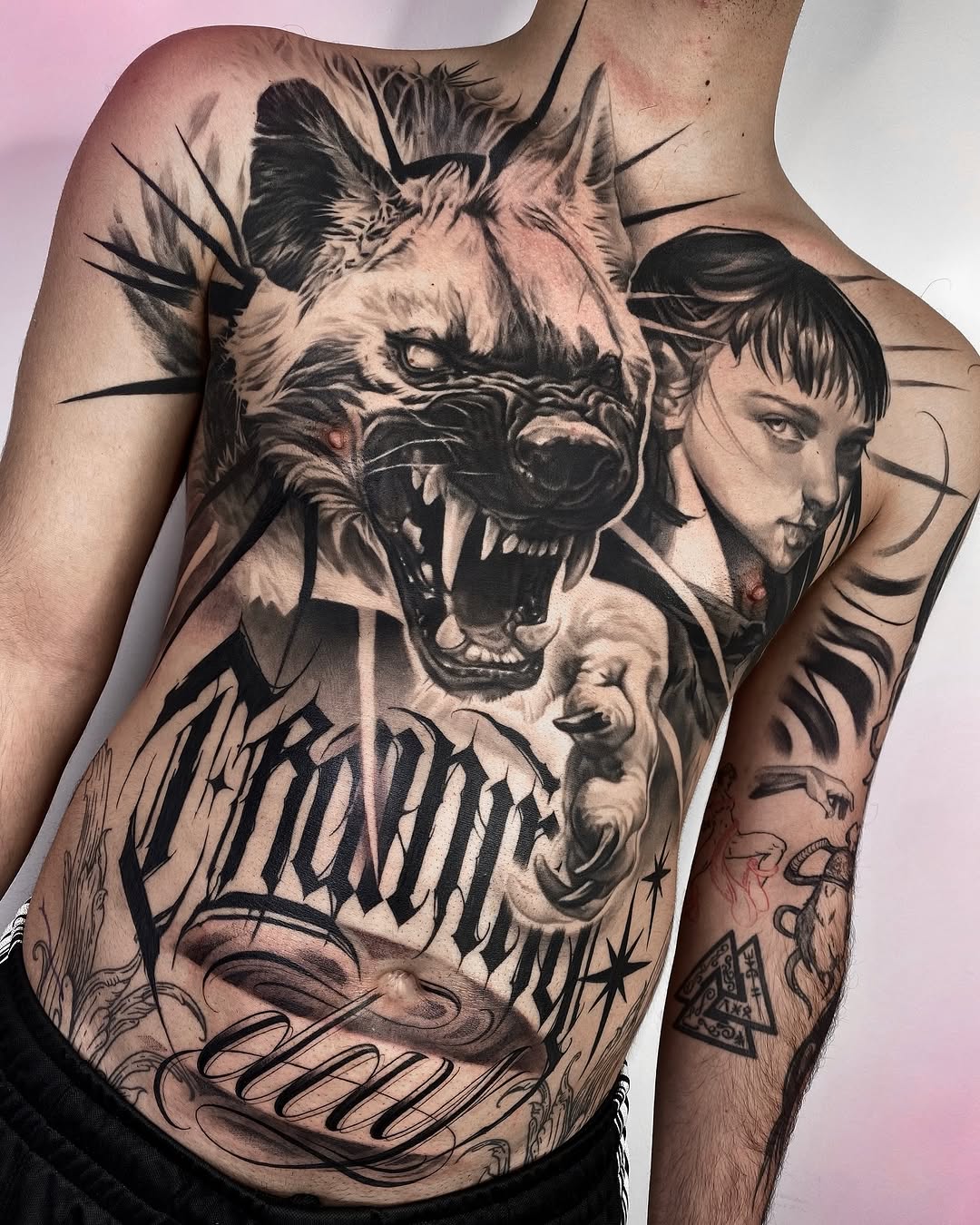 Large black and grey chest tattoo featuring a roaring lion face with human faces integrated into the design, surrounded by decorative text and line work.