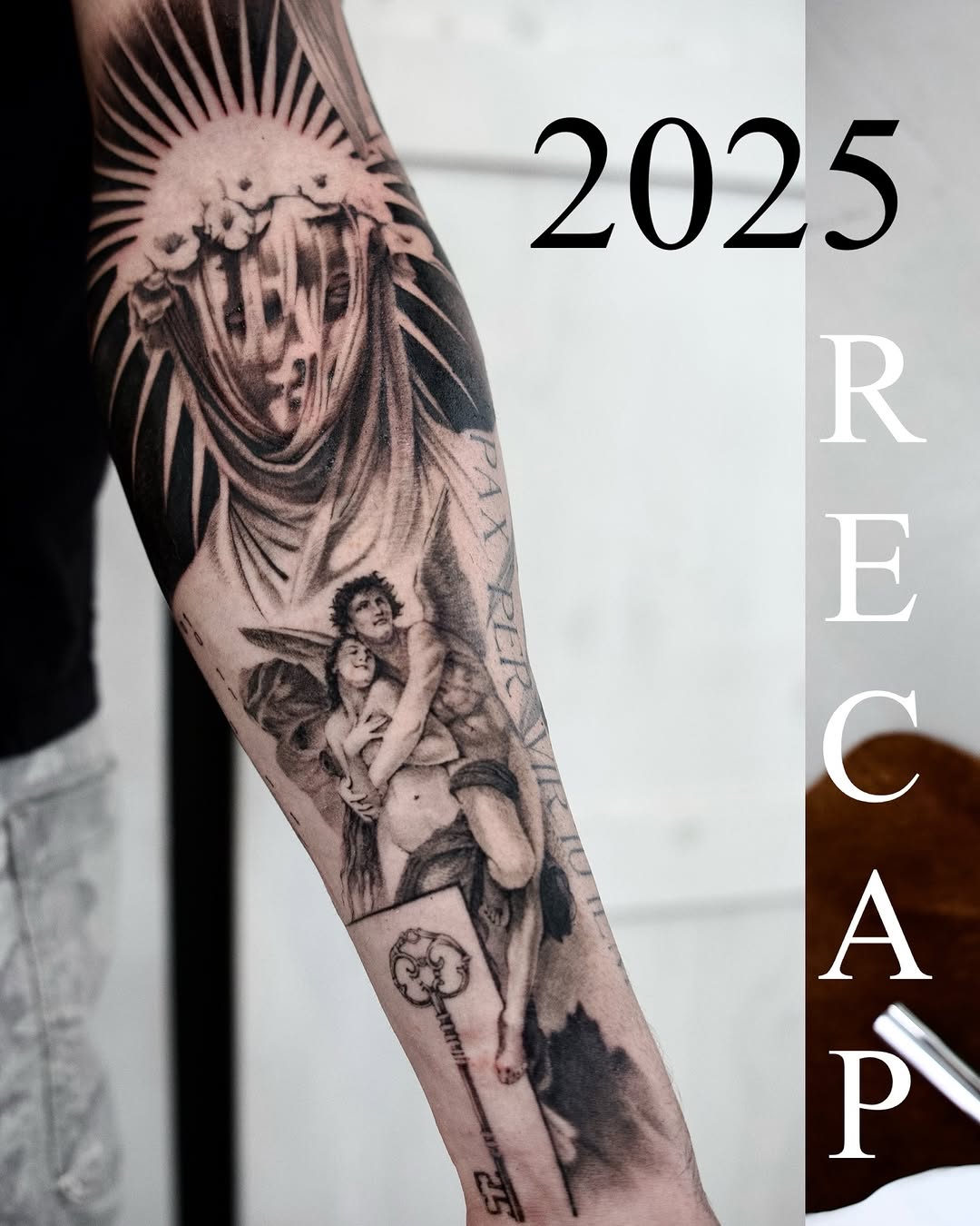 Black and grey arm tattoo featuring a winged angelic figure with a halo above smaller cherub figures, displayed on inner forearm.