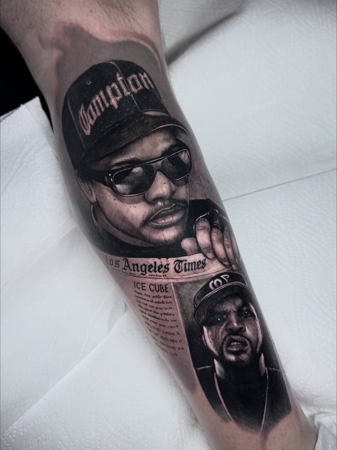 Black and grey portrait tattoo on forearm featuring two men wearing sunglasses and caps with Los Angeles Times newspaper clipping below.