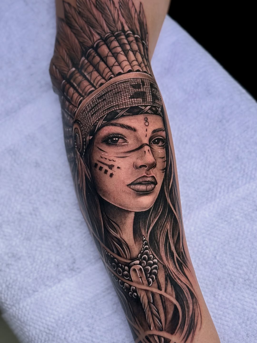 Indigenous woman portrait with feathered headdress, facial markings, and beaded necklace on forearm.