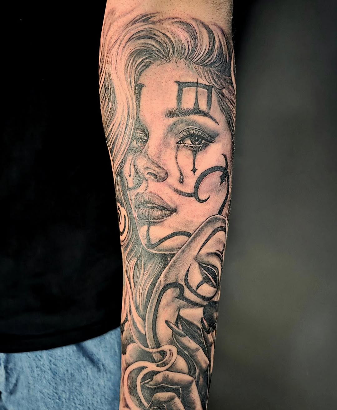 Woman's face with wavy hair, tears, geometric forehead symbol, and crescent moon facial markings tattooed on forearm in black and grey.