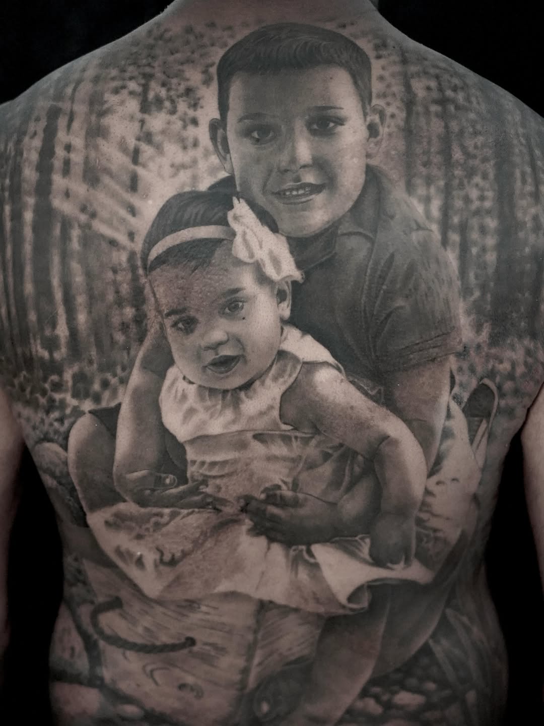 Black and grey photorealistic portrait tattoo of two young children on upper back, rendered with monochromatic tonal blending.