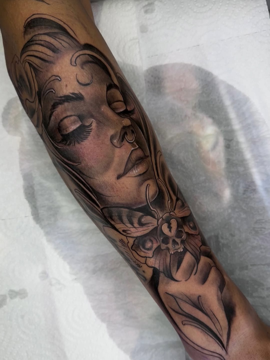 Detailed black and grey portrait tattoo of a man's face with ornamental flourishes on the forearm, featuring smooth shading and realistic facial features.