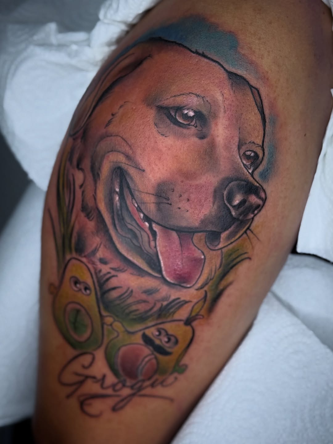 Dog's head portrait with tongue out in black and grey shading on upper arm, with decorative dice and scrollwork below.
