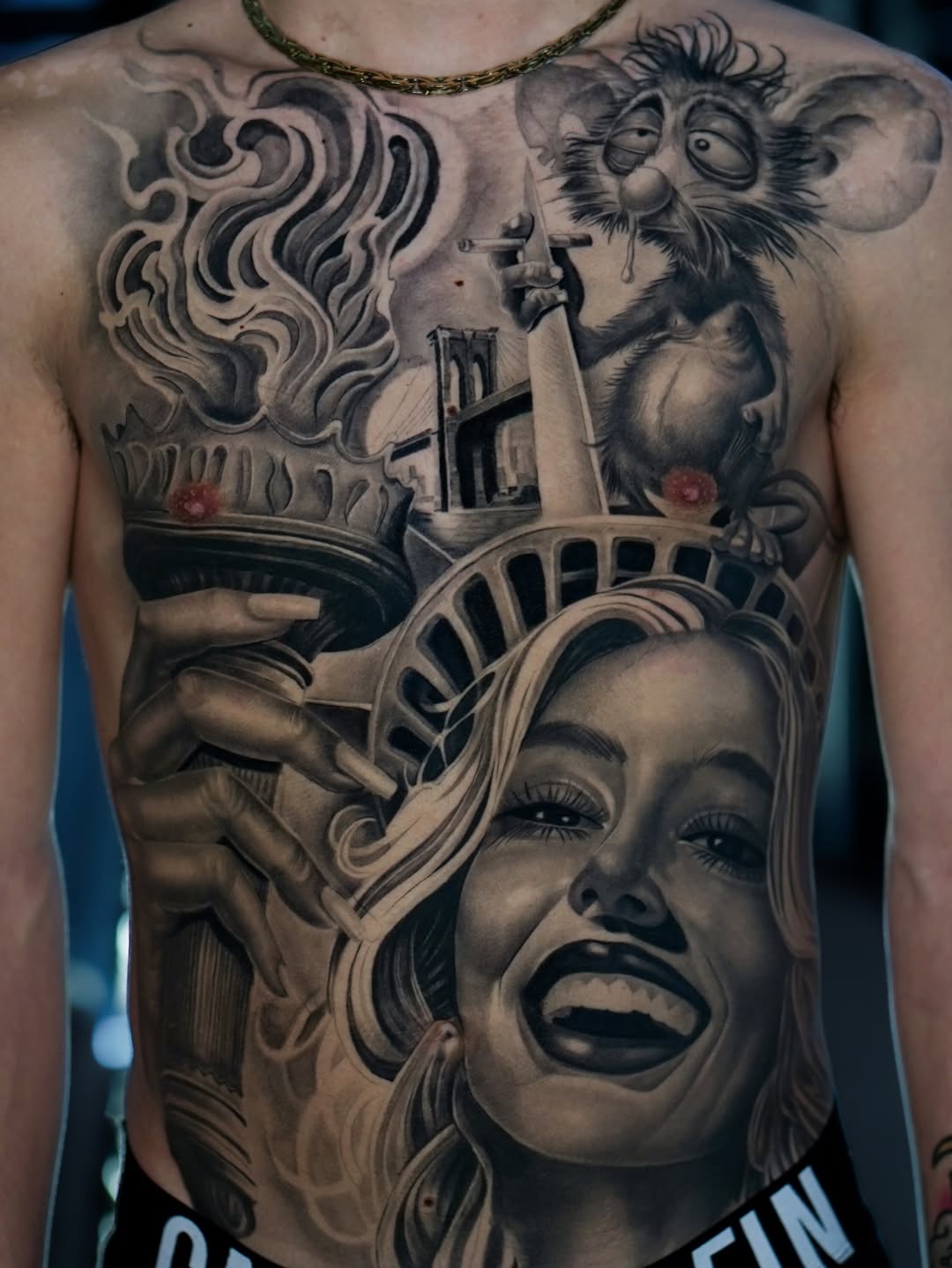 Chest tattoo featuring a woman with Statue of Liberty crown, playful monkey, ornate flames, and architectural elements in black and grey shading.