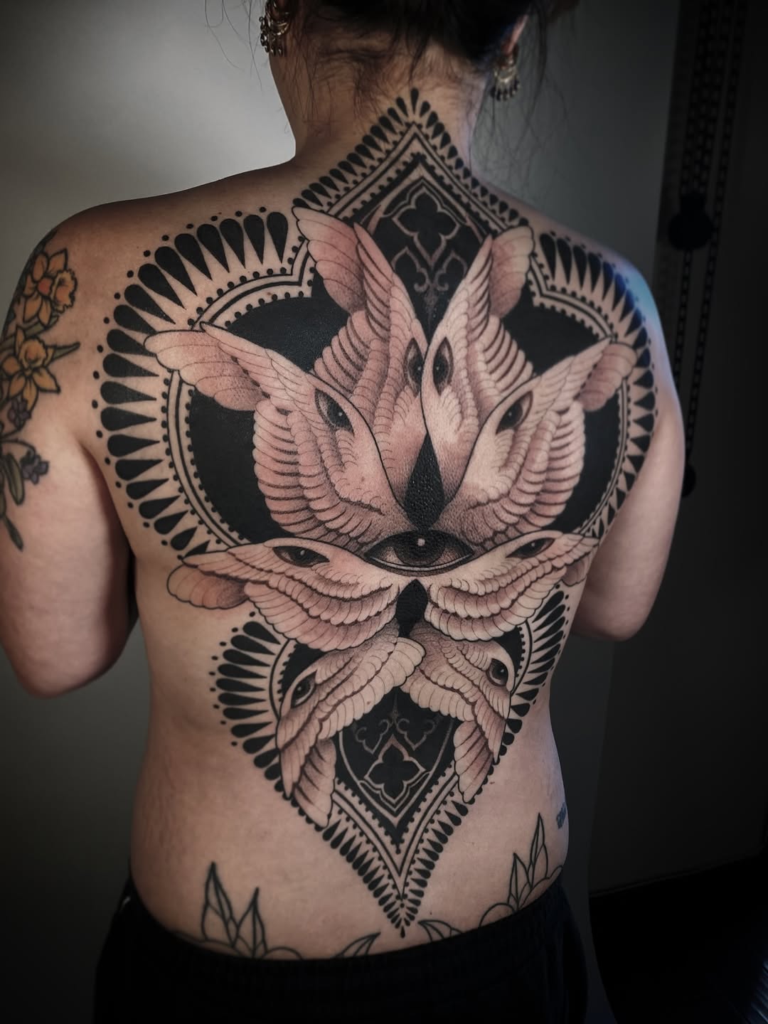 Back tattoo featuring four winged birds with detailed feather work arranged around geometric diamond shapes, rendered in black and grey with soft shading.