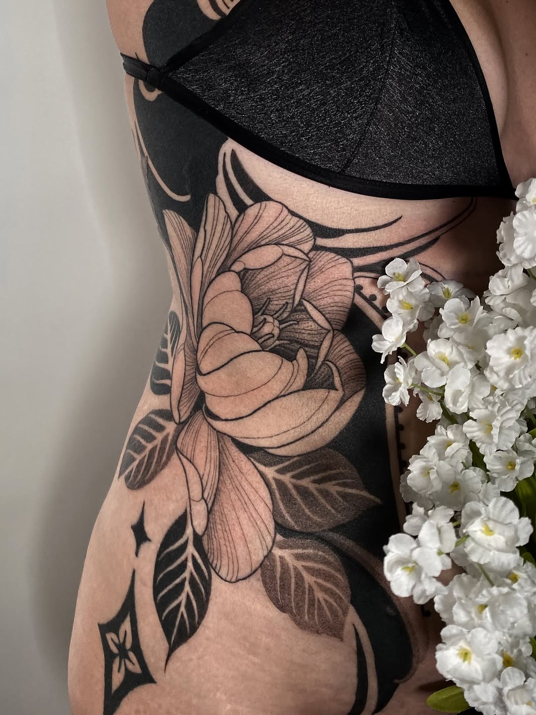 Black and grey floral tattoo featuring a detailed peony with layered petals and ornamental leaves on the ribs and torso.