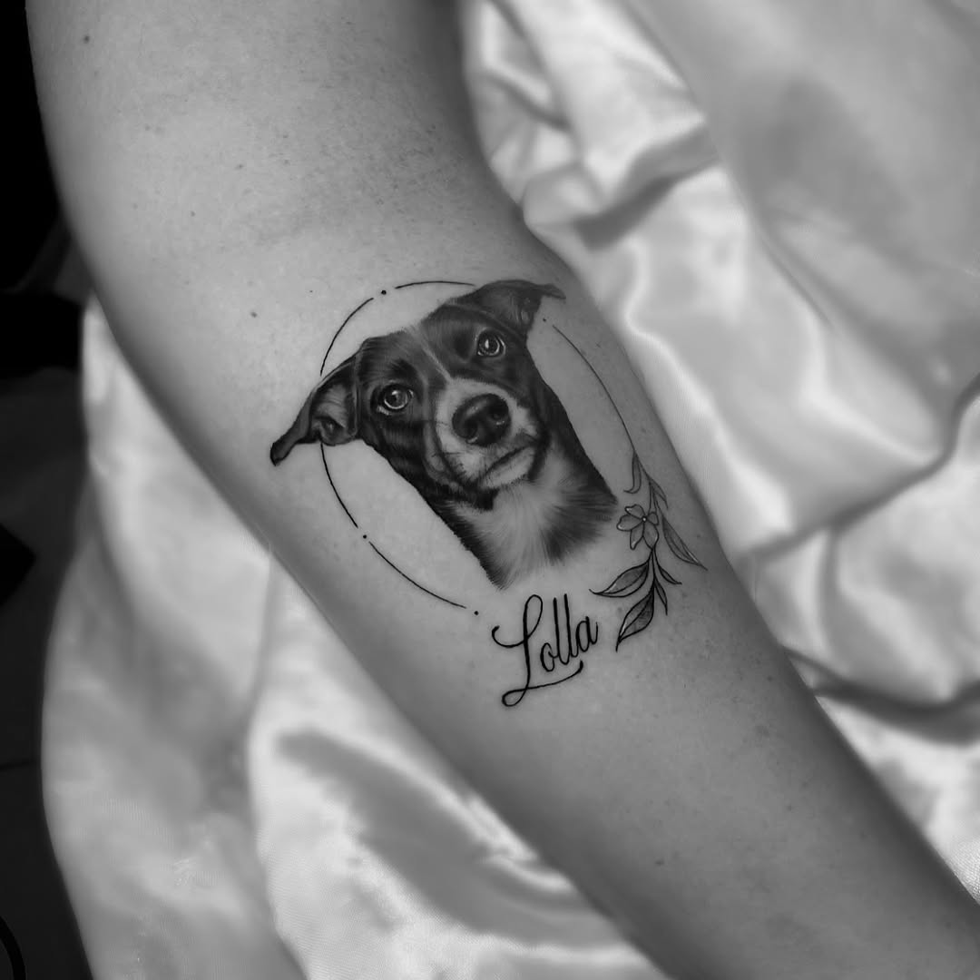 Black and grey portrait of a dog's face with tilted head, enclosed in circular line frame with floral sprigs and script name below on arm.