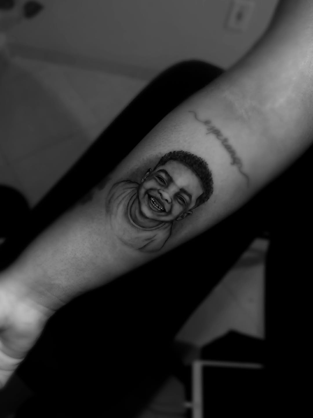 Portrait tattoo of a smiling young man's face on the inner forearm, rendered in black and grey with monochromatic tonal blending.