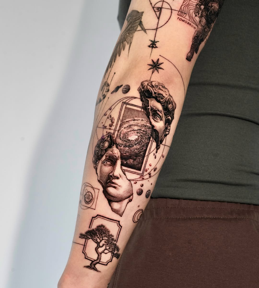 Surreal black and grey forearm tattoo featuring classical busts, celestial elements, and organic forms with soft gradient shading.
