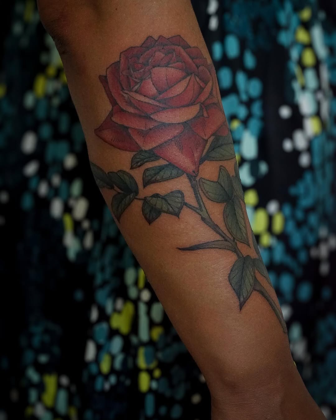 Red rose with green leaves and stem on inner forearm, rendered in black and grey with soft shading and realism.