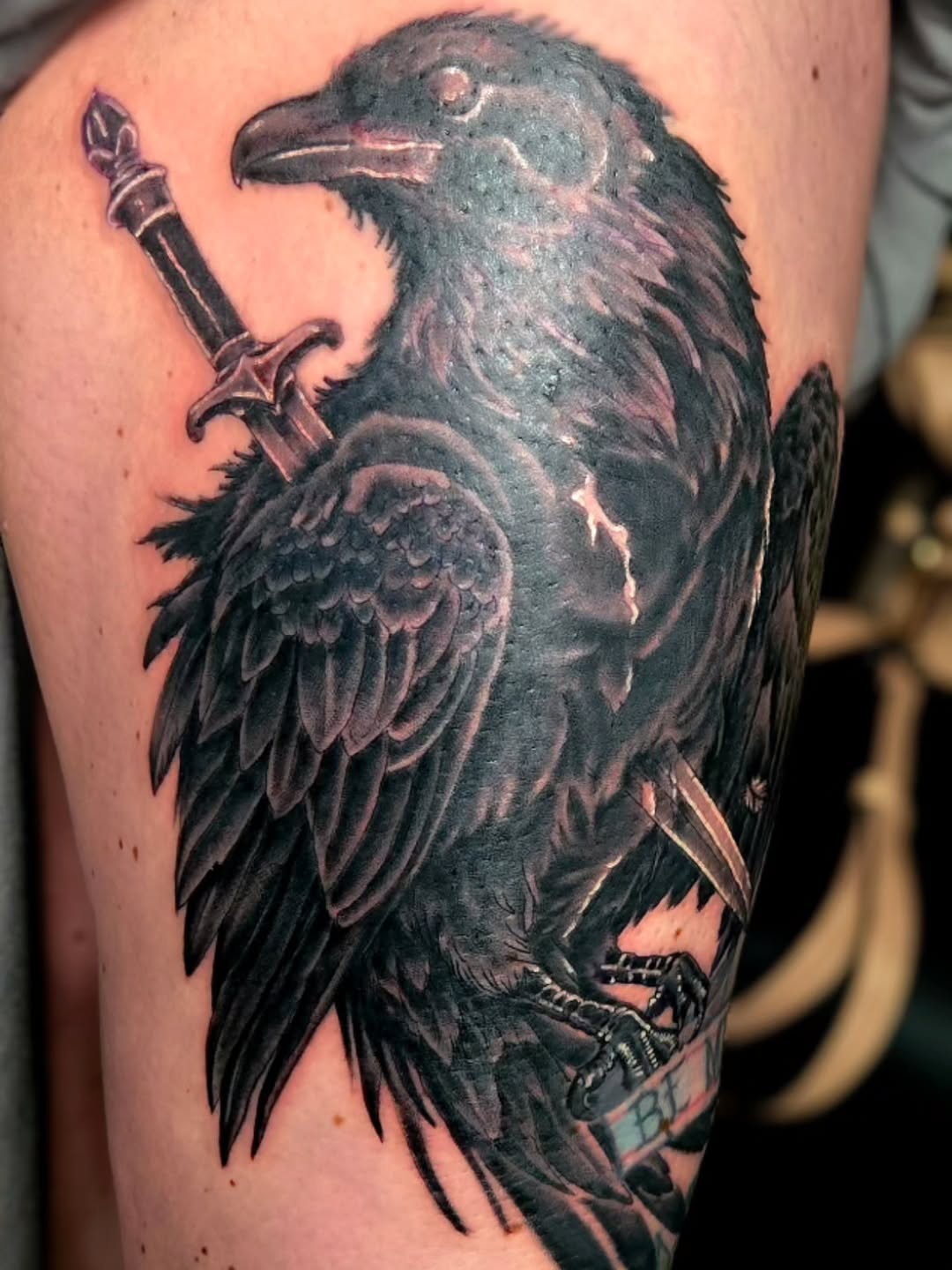 Black and grey raven tattoo on upper arm holding a dagger and banner, detailed feather shading with realistic highlights.