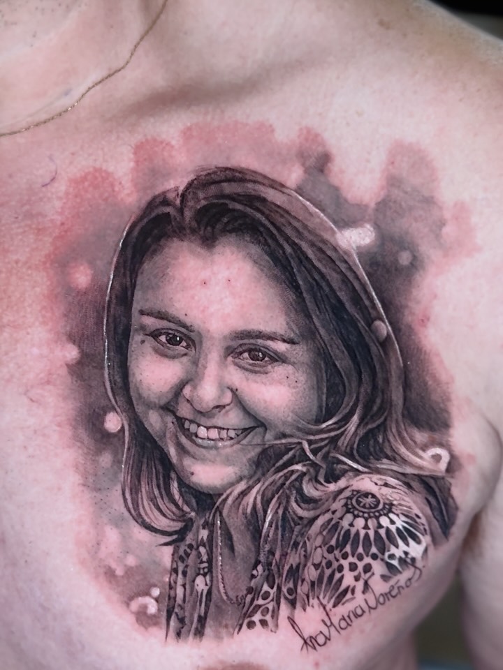 Portrait tattoo of a smiling woman with long hair and decorative shoulder piece in black and grey on chest.