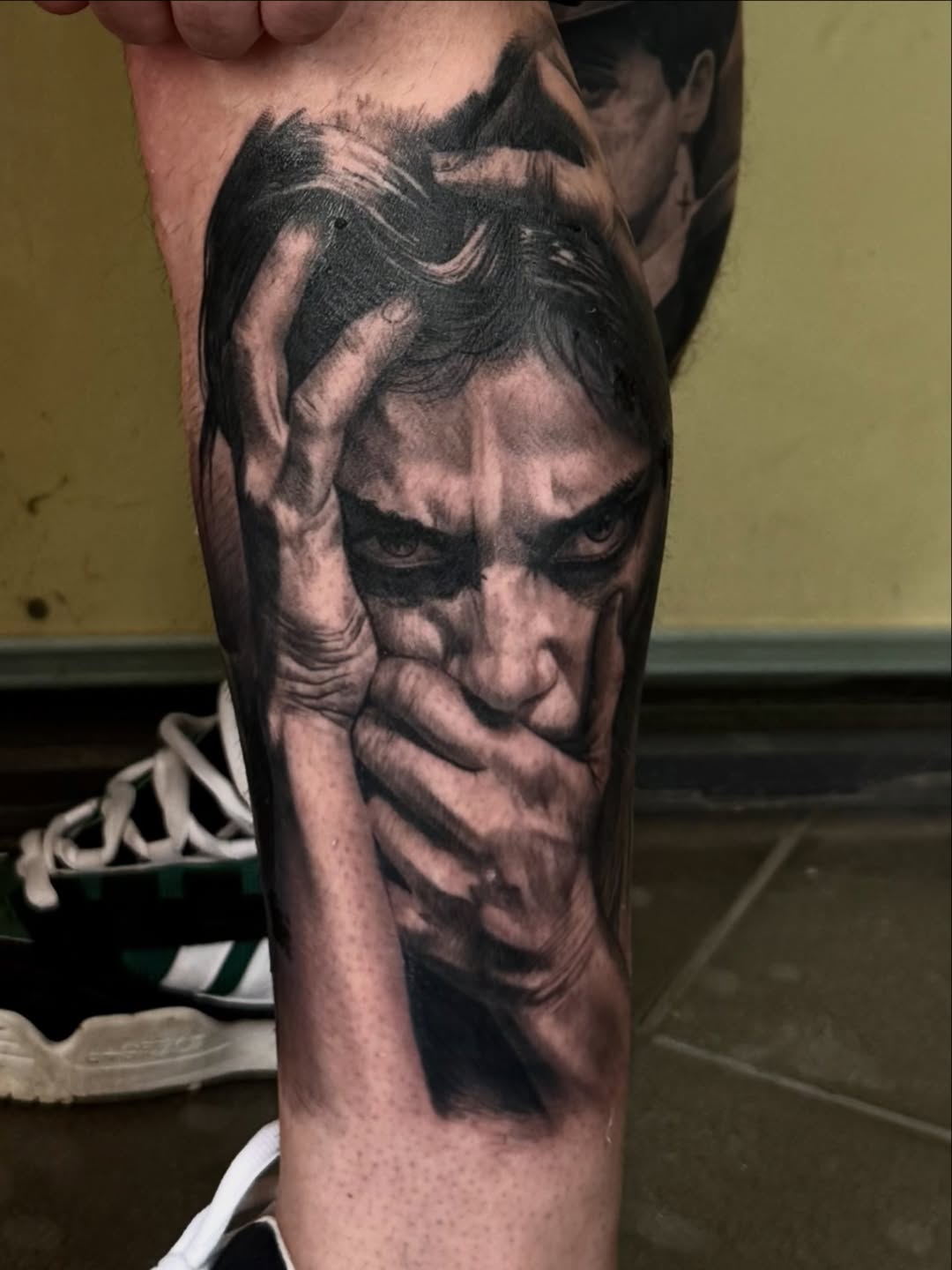 Black and grey portrait tattoo of a man's face with dramatic shading on the lower leg, showing detailed facial features and emotional expression.