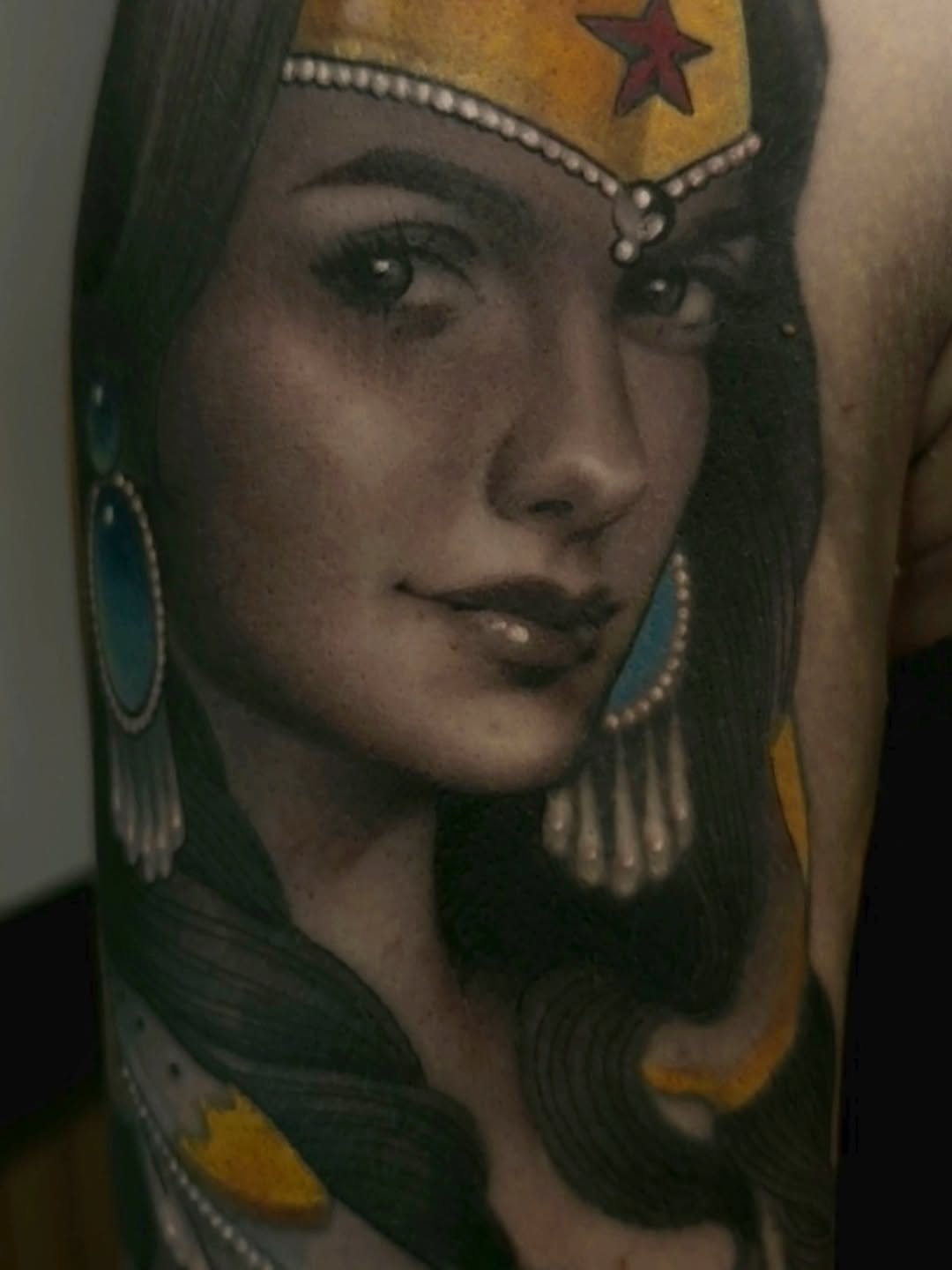 Portrait tattoo of a woman with long dark hair, gold headpiece with red star, pearl necklace and earrings, rendered in black and grey on arm.