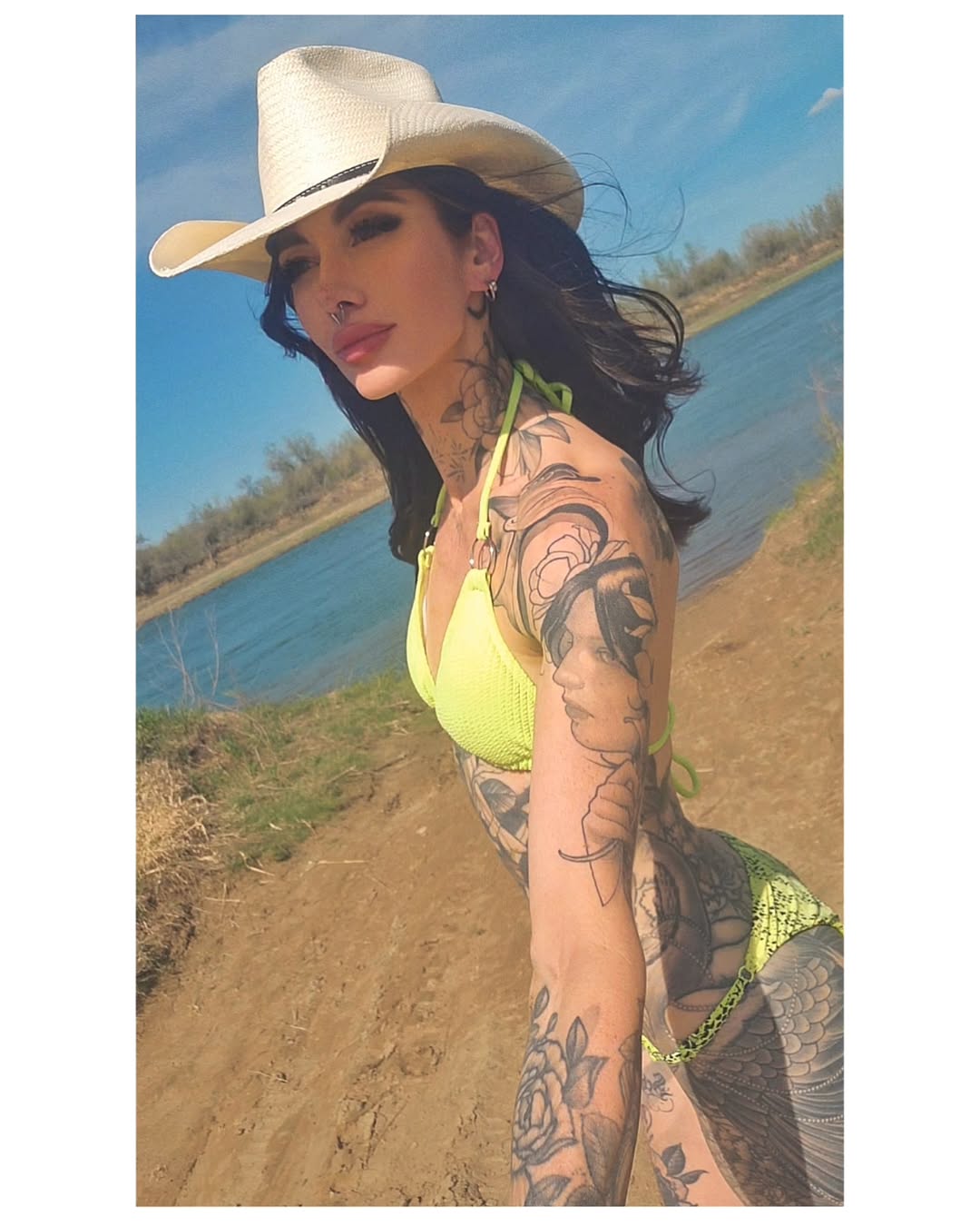 Woman's arm and torso featuring black and grey portrait and decorative tattoos, shown outdoors by water wearing a cowboy hat.