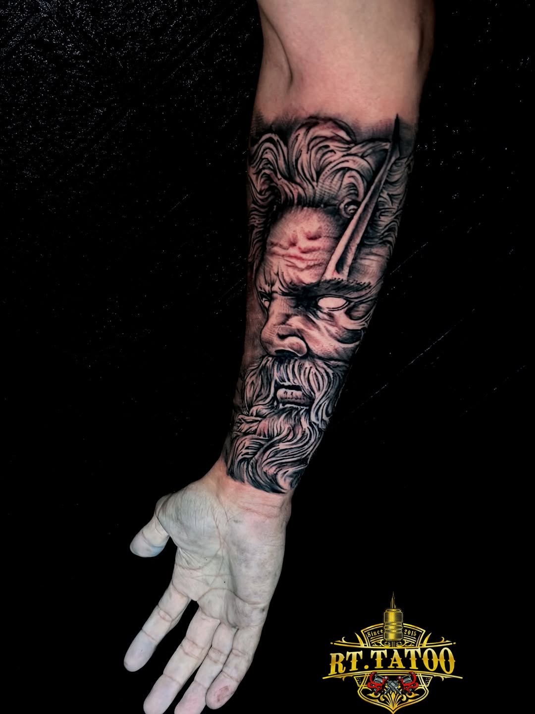 Stacked portrait tattoo featuring a bearded man's face above a cherub or angel head, rendered in black and grey on the forearm.