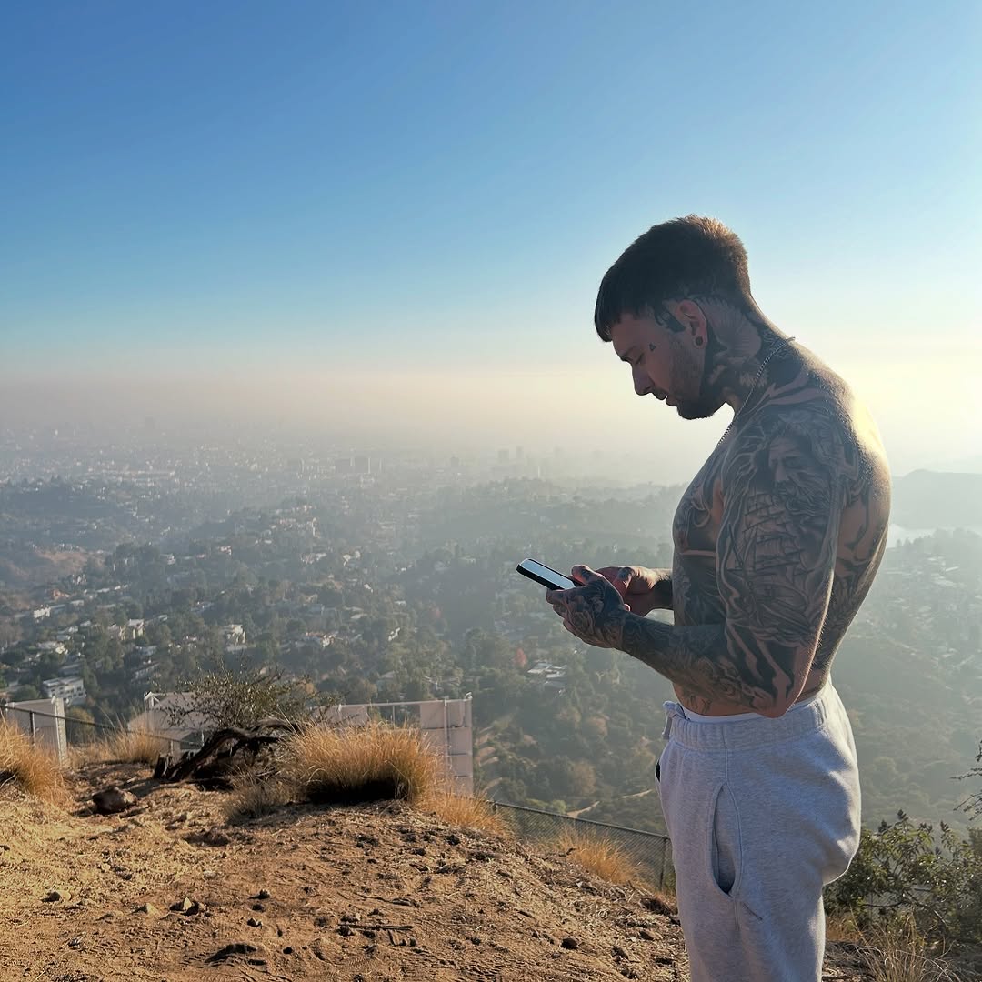 Man with extensive black and grey back and chest tattoo featuring detailed imagery, shirtless outdoors overlooking a cityscape valley.