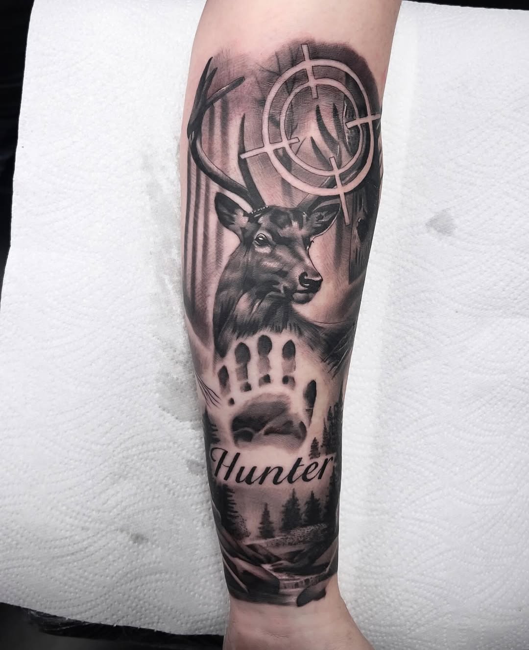 Black and grey sleeve tattoo featuring a detailed deer head with antlers, hunting rifle crosshair, paw print, forest landscape, and 'Hunter' text on forearm.