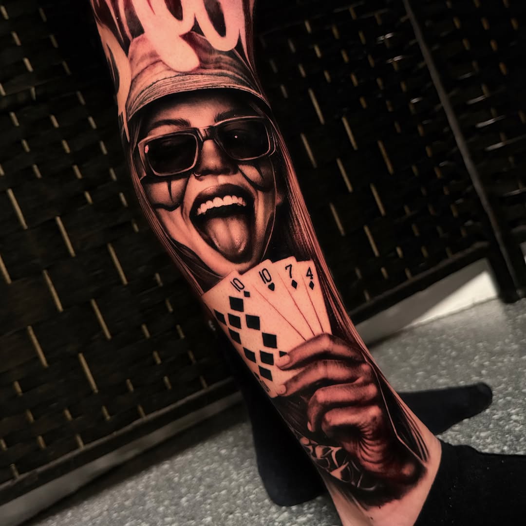 Black and grey portrait tattoo of a person wearing sunglasses and a tall hat, holding playing cards on the forearm.