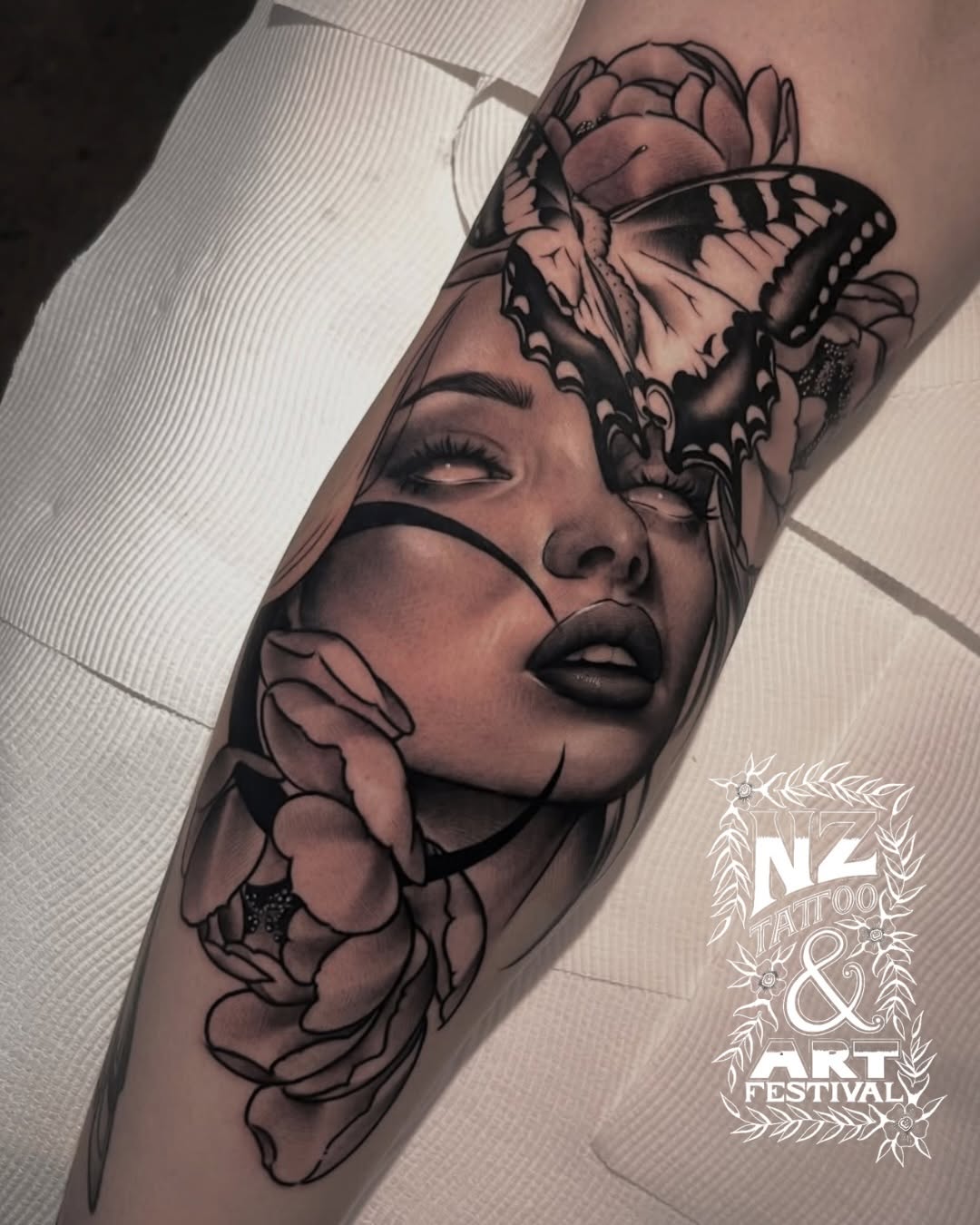 Black and grey portrait tattoo of a woman's face with closed eyes, surrounded by roses and a butterfly on the forearm.