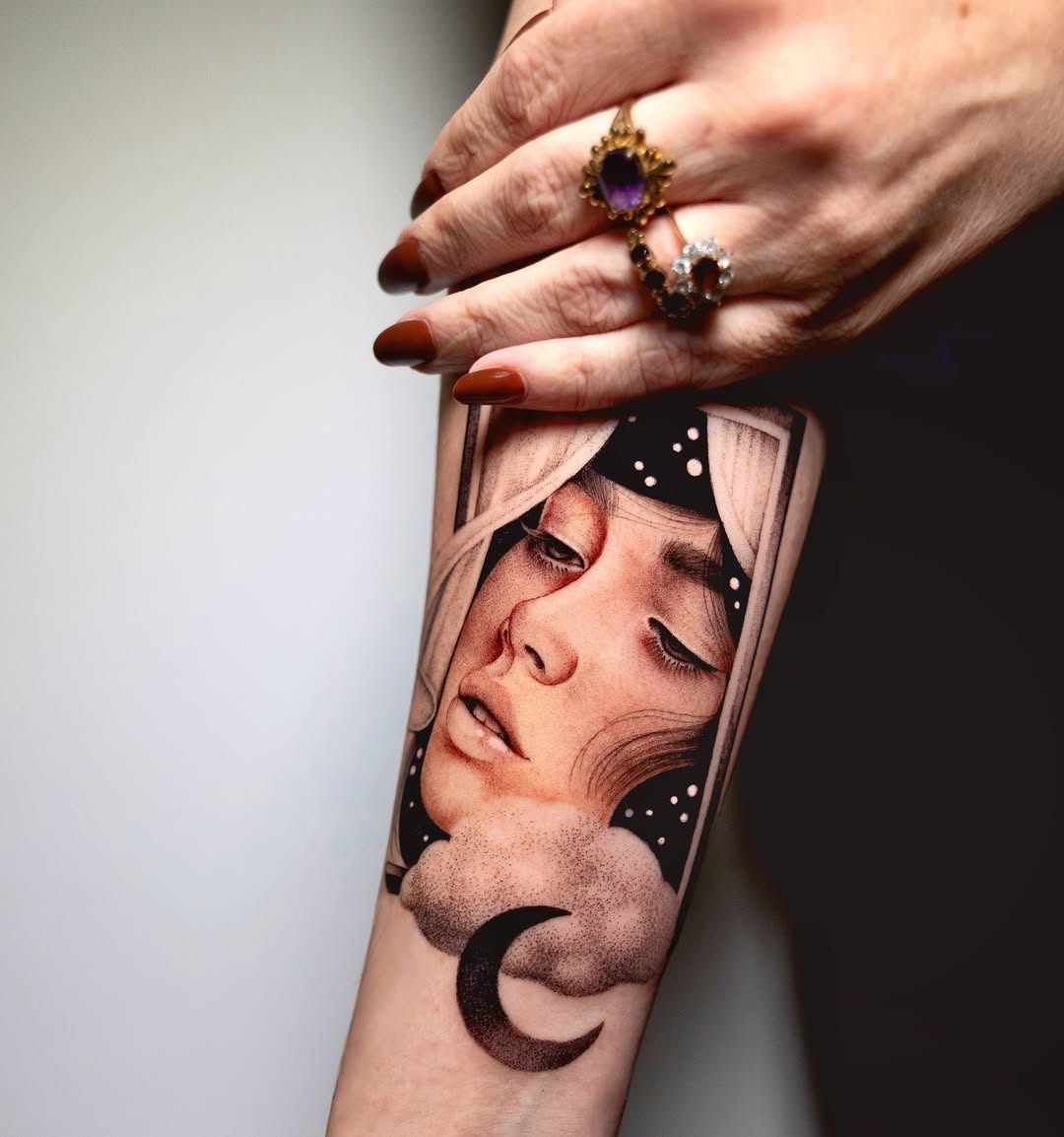 Portrait tattoo of a person's face with closed eyes and crescent moon below, decorated with stars, in black and grey on forearm.