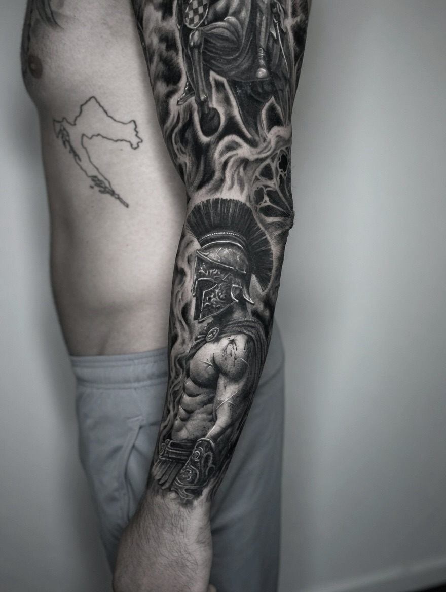 Black and grey sleeve tattoo of a Spartan warrior with plumed helmet, flames, and a soldier figure covering the entire arm.