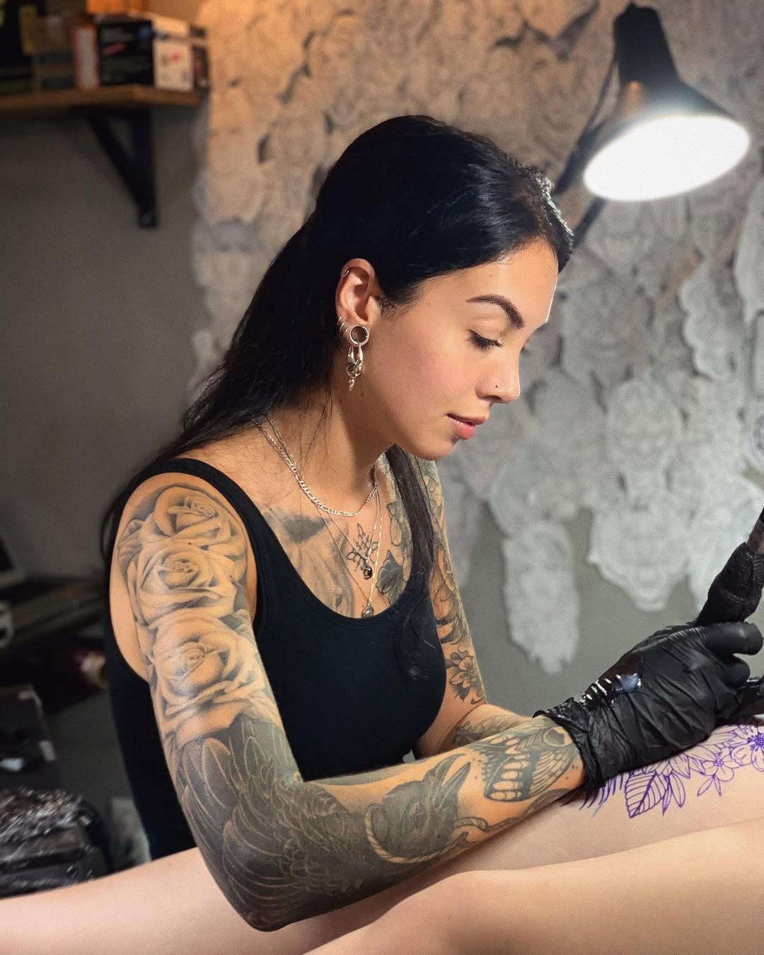 Tattoo artist applying black and grey floral design with purple accents to client's forearm under studio lighting.