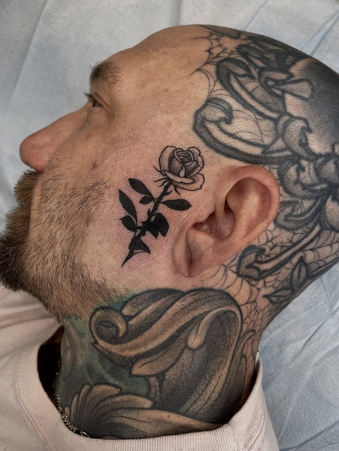 Black and grey rose tattoo on man's cheek near ear with decorative leaves and stem detail.