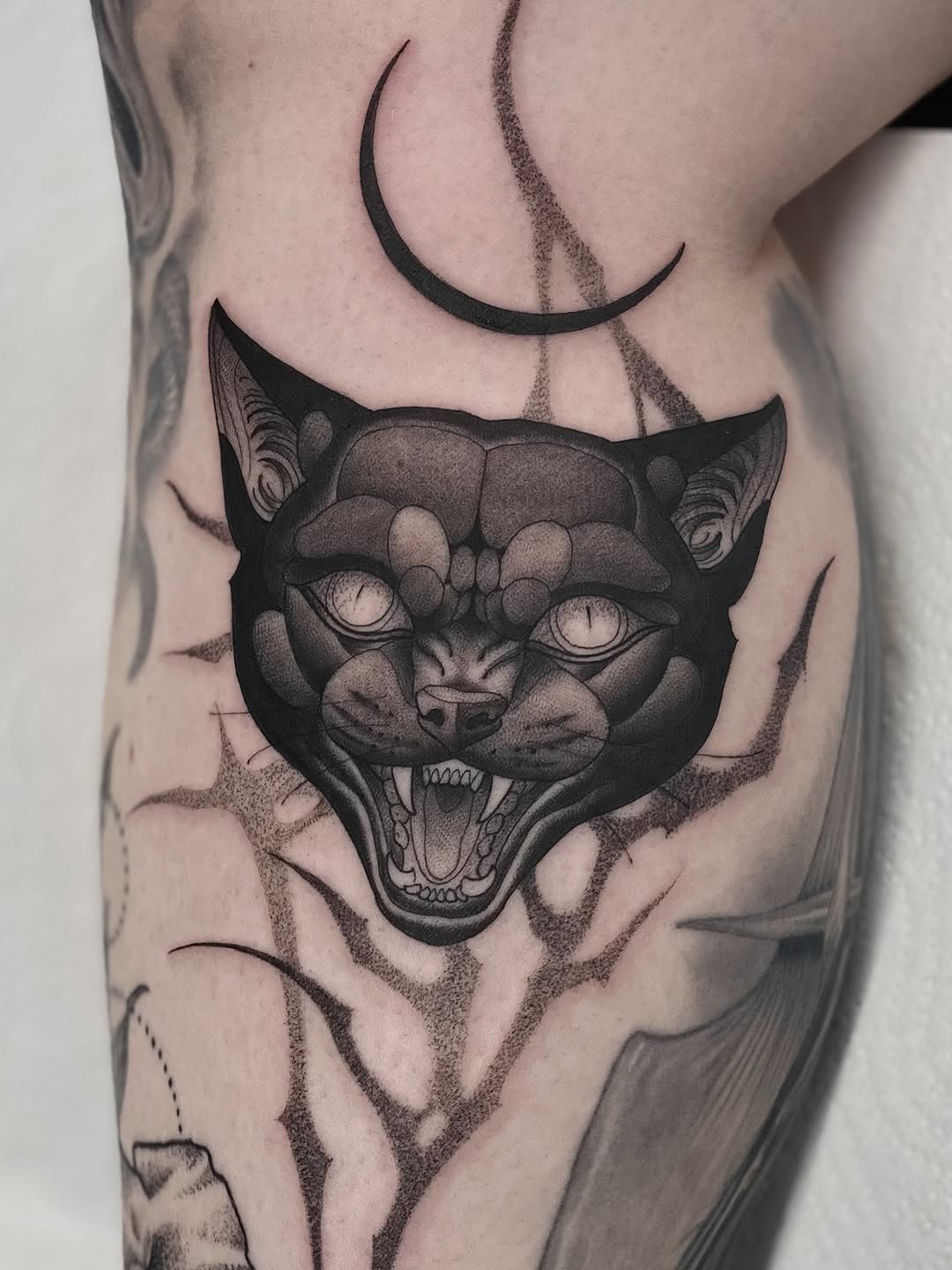 Demonic cat head with bulging eyes and bared teeth, centered on thigh with crescent moons above, rendered in black and grey with smooth shading.