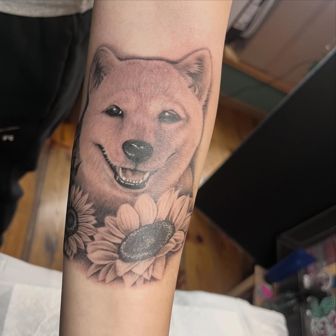 Smiling dog head with sunflowers on forearm, rendered in black and grey with soft shading and realistic detail.