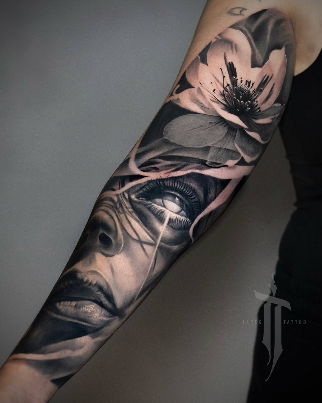 Black and grey sleeve tattoo featuring a woman's face with closed eyes and a delicate flower blooming from her head, with smooth shading and realistic details on the forearm.
