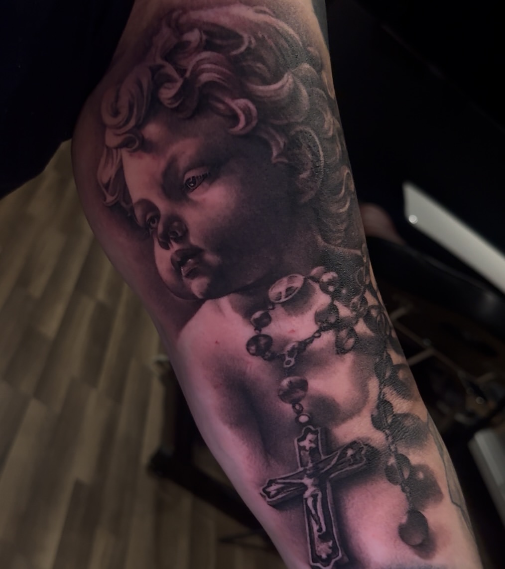 Cherub face with curled hair and rosary beads tattooed on upper arm in black and grey with soft shading.