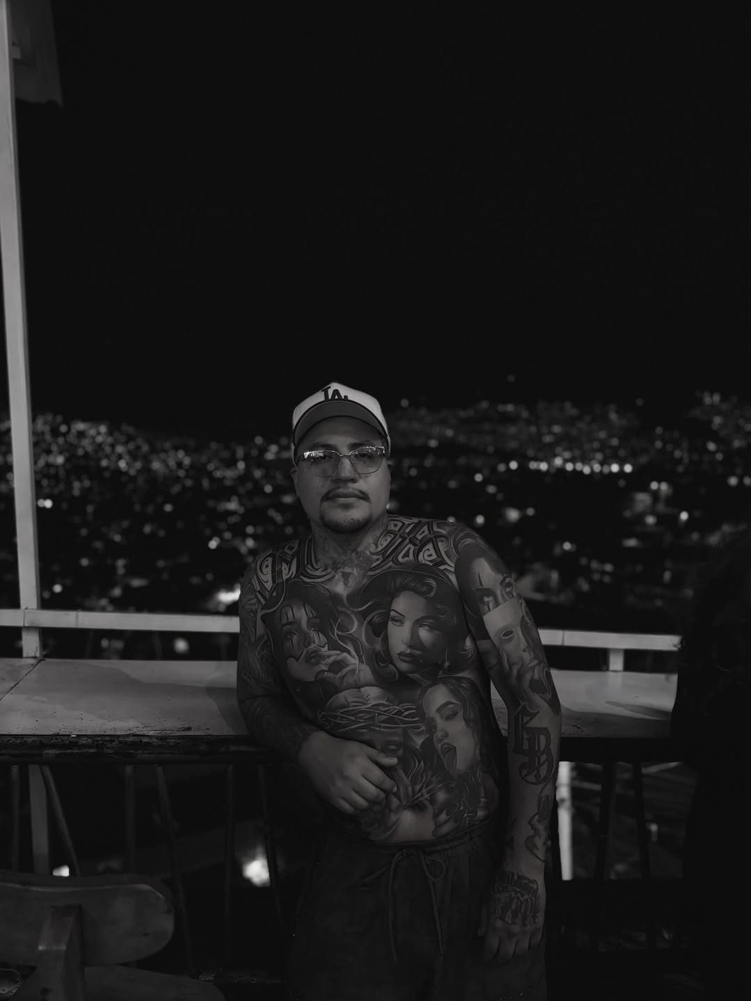Black and grey portrait tattoos covering a man's chest and arm, featuring multiple faces with detailed shading against a city skyline background.