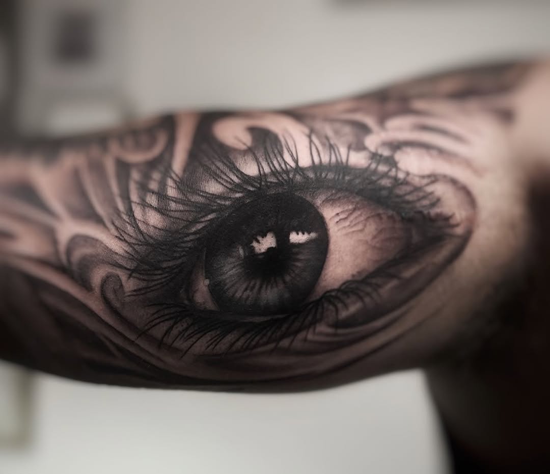 Realistic black and grey eye tattoo with detailed iris, lashes, and ornamental linework on inner forearm showing smooth shading gradients.