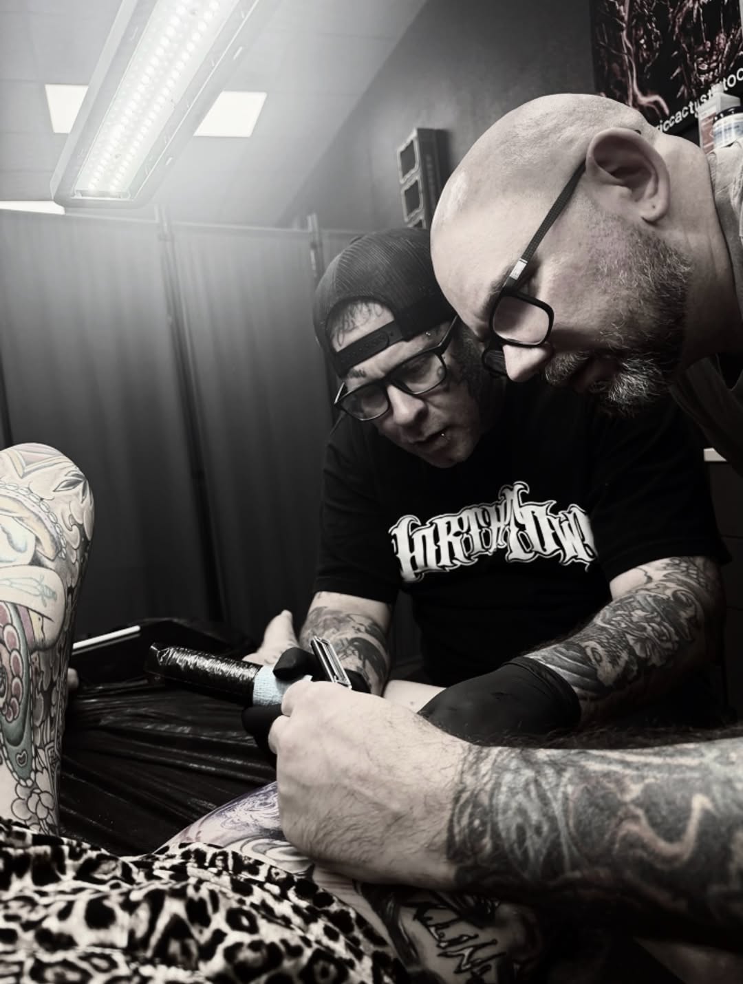 Tattoo artist with glasses applies black and grey ink to client's tattooed arm in bright studio setting.
