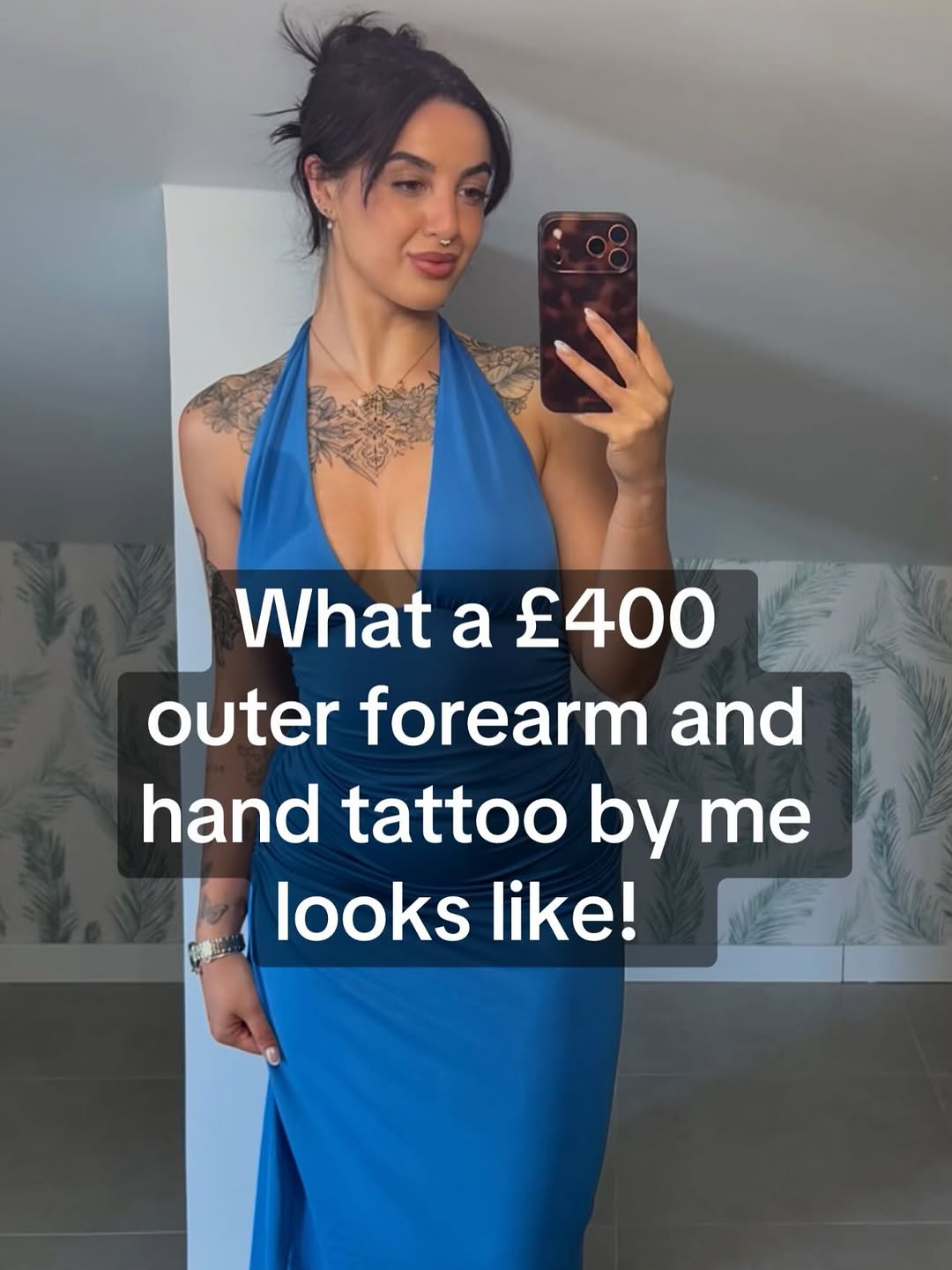 Intricate black and grey chest and outer forearm tattoo with ornamental patterns and detailed linework on a woman in a blue halter dress.