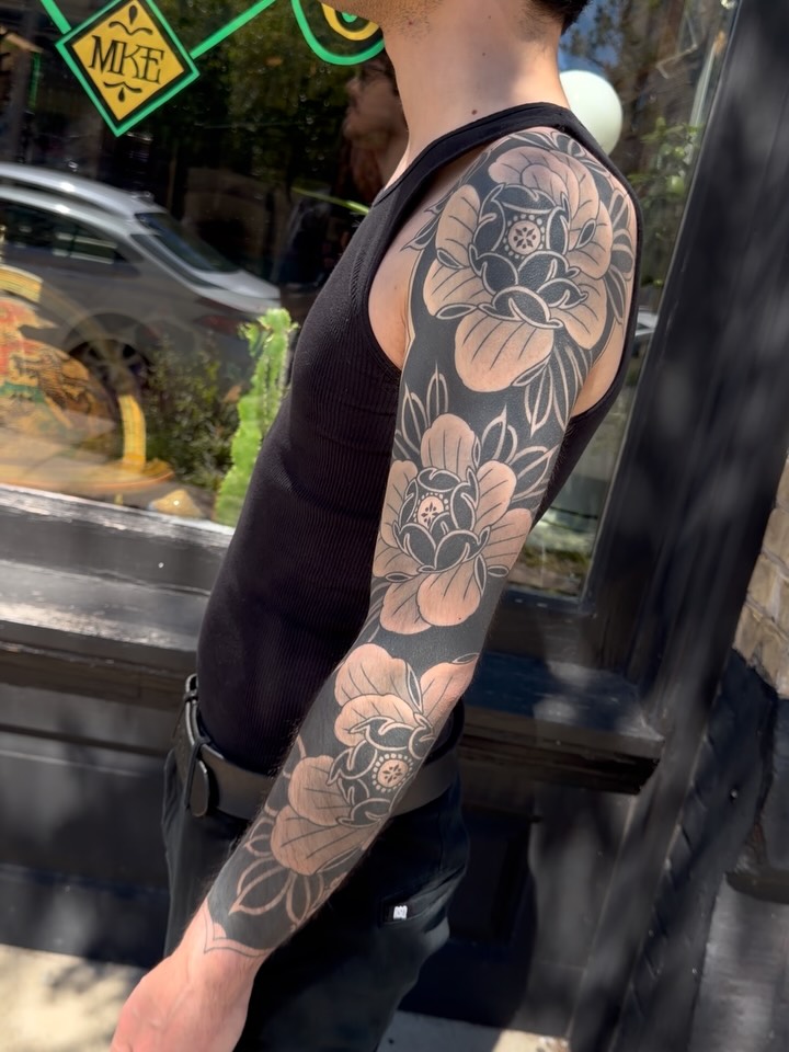 Full arm sleeve tattoo featuring multiple large peony flowers with detailed shading in black and grey on the shoulder and forearm.