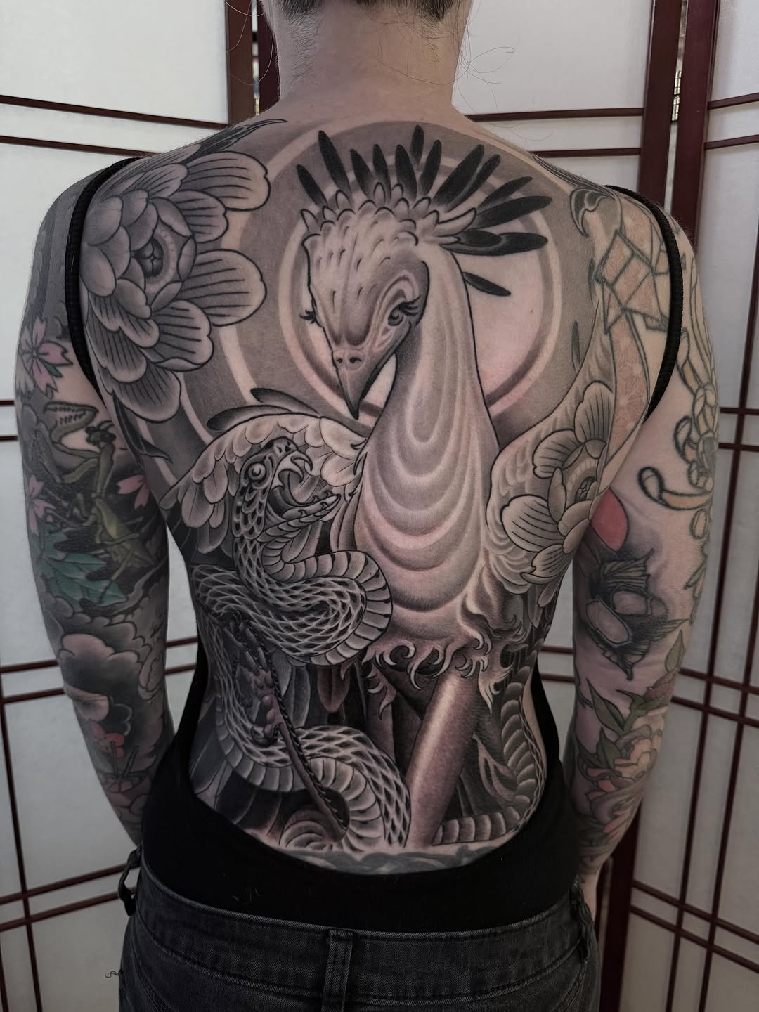 Large back tattoo featuring a crane with spread wings and decorative feathers in black and grey with soft shading.