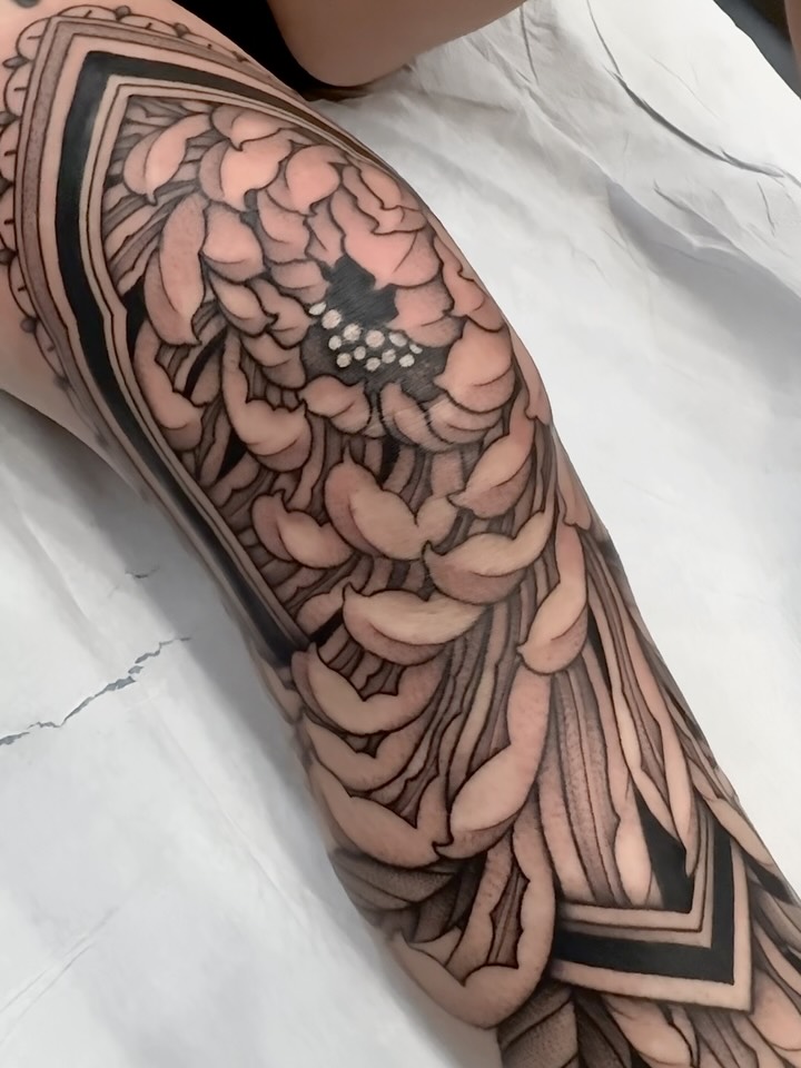 Large intricate peony flower tattoo on upper arm with layered petals, fine line work, and a small bird detail, rendered in black and grey shading.