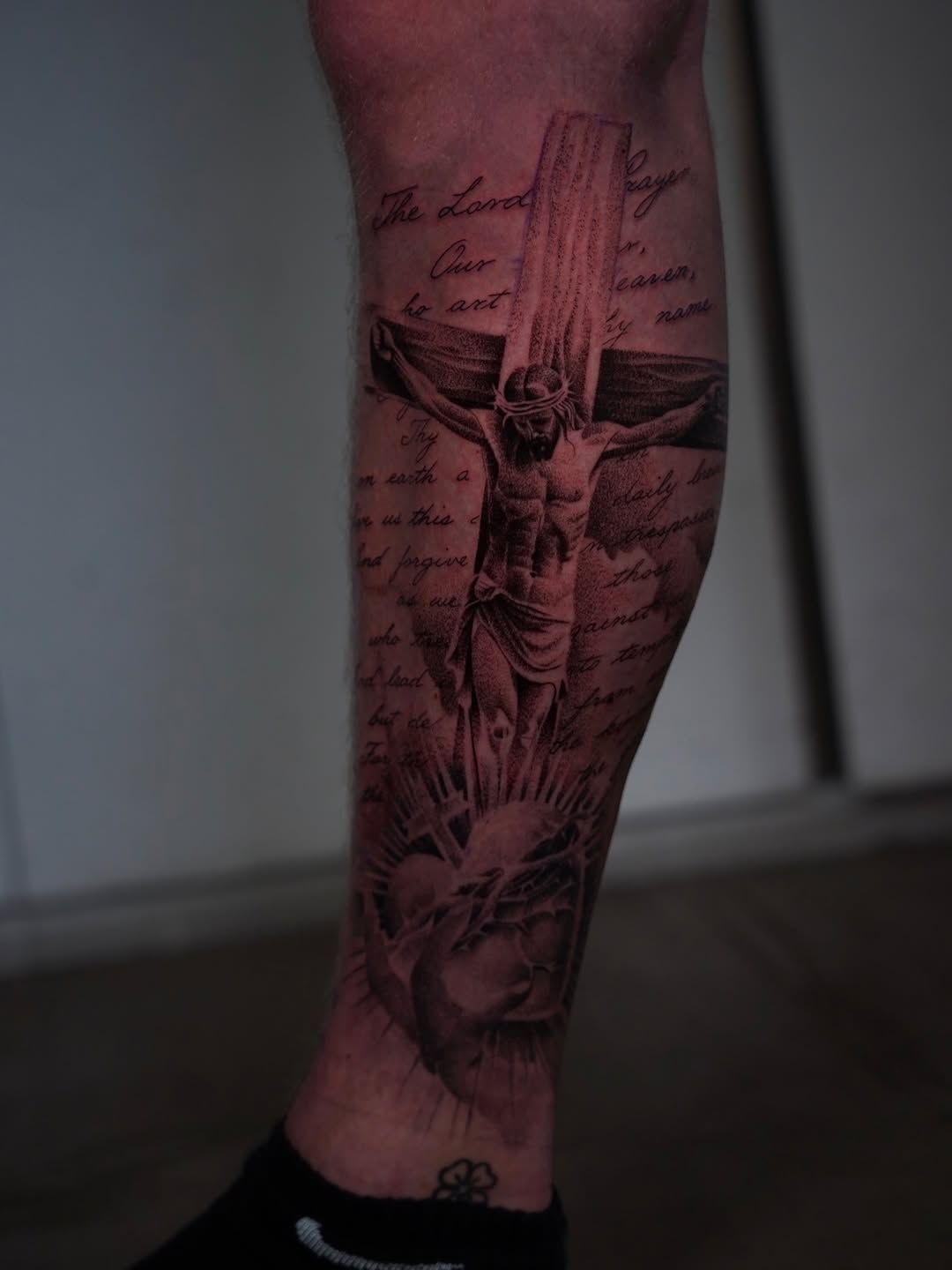 Crucifix tattoo with flowing fabric and radiating light on calf, surrounded by handwritten script text in black and grey.