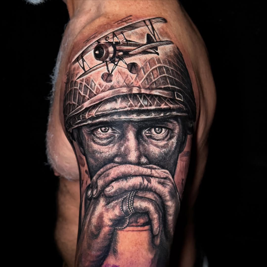 Bearded man's face with ornate headpiece and biplane above, rendered in black and grey realism on upper arm.