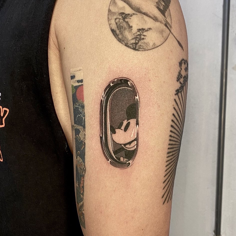 Mickey Mouse portrait in an oval chrome frame on upper arm, rendered in black and grey with smooth shading and highlights.