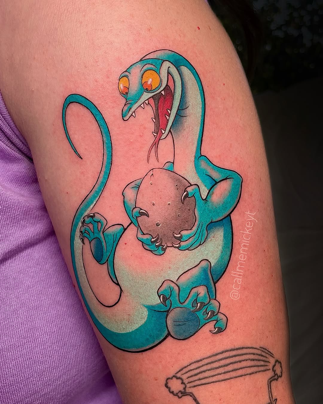 A colorful cartoon sea creature with an angry expression holding a round character, tattooed on an upper arm.