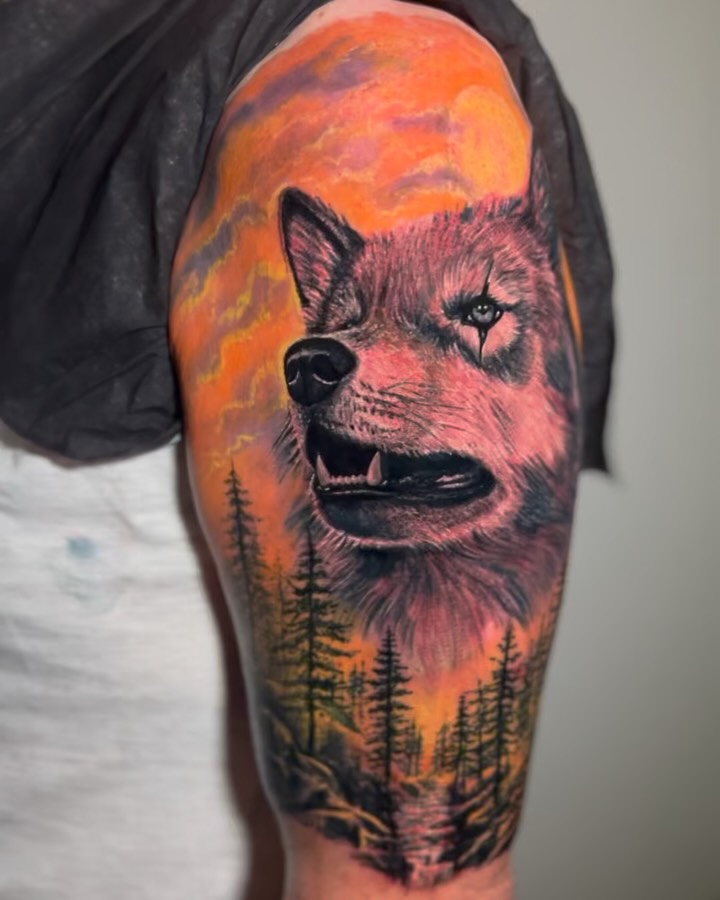 Wolf head with open mouth against orange sunset sky and dark forest silhouettes on upper arm.