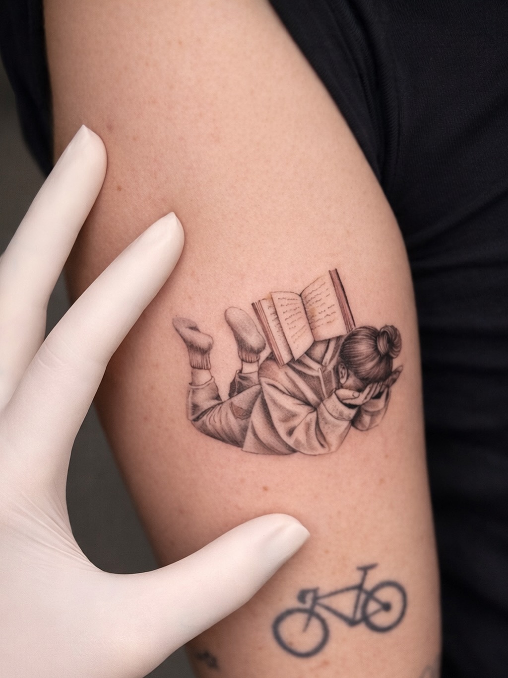 A girl reading an open book with legs up, rendered in fine monochromatic black and grey linework on the upper arm.