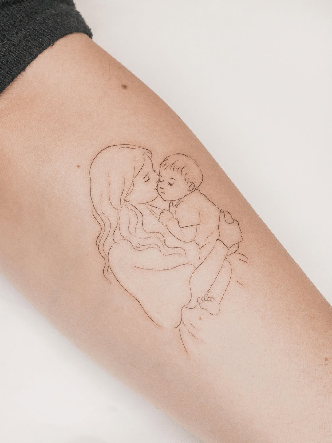 A mother holding a baby in her arms, rendered in delicate thin-line sketch style on the upper arm.