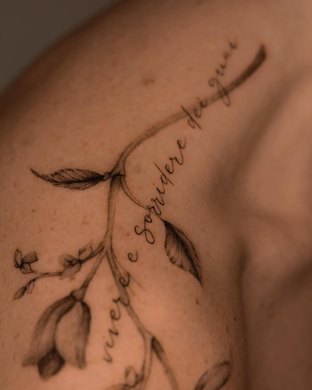 Delicate botanical tattoo with feathered leaves and cursive script text flowing along branches on shoulder area.