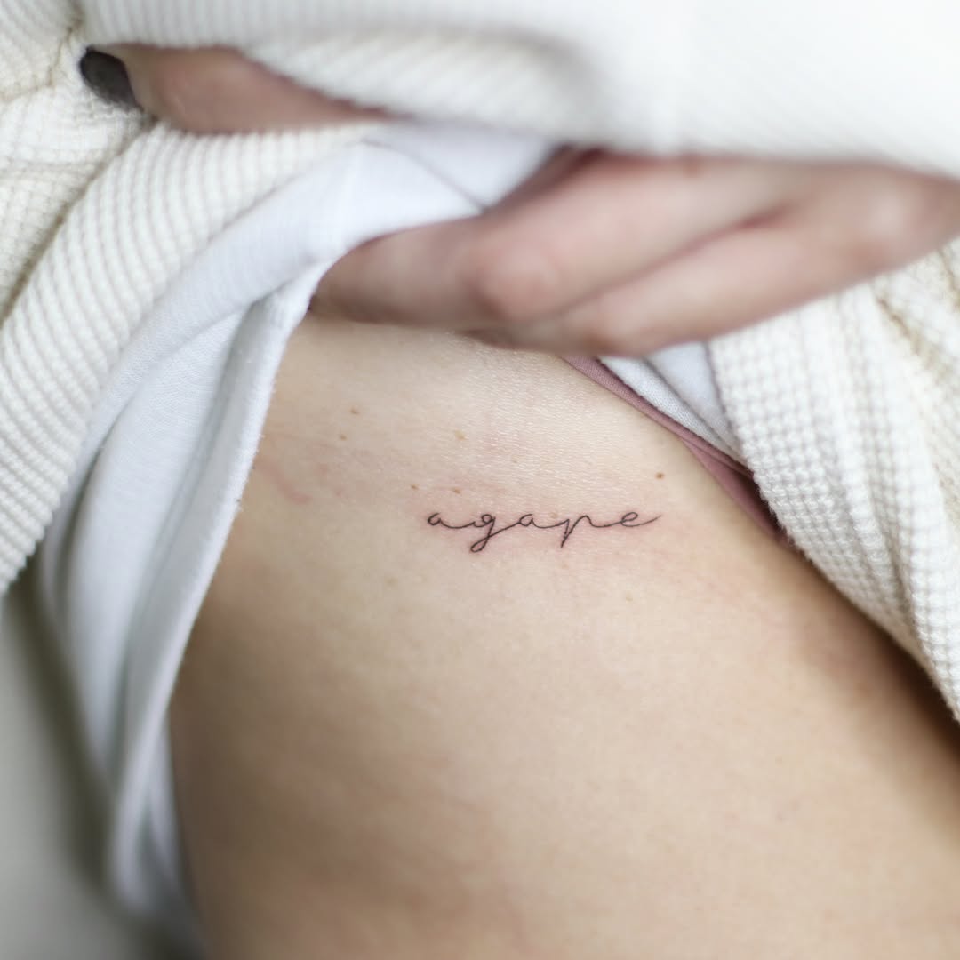 Delicate cursive script reading 'agape' in thin linework on the upper arm or shoulder area.